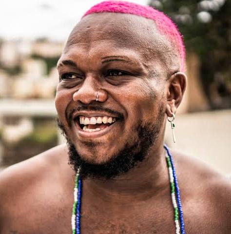 Homem de cabelo rosa (rapper Djonga) sorrindo.