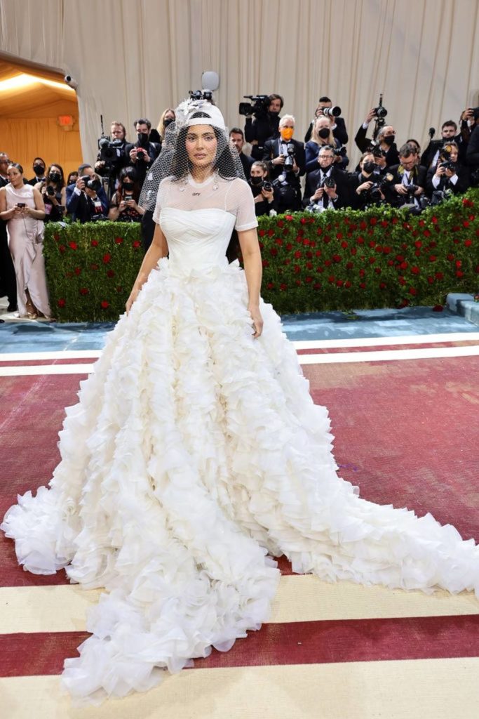 A imagem tem ao centro Kylie Jenner usando um vestido branco de noiva com um chapéu branco e uma espécie de véu