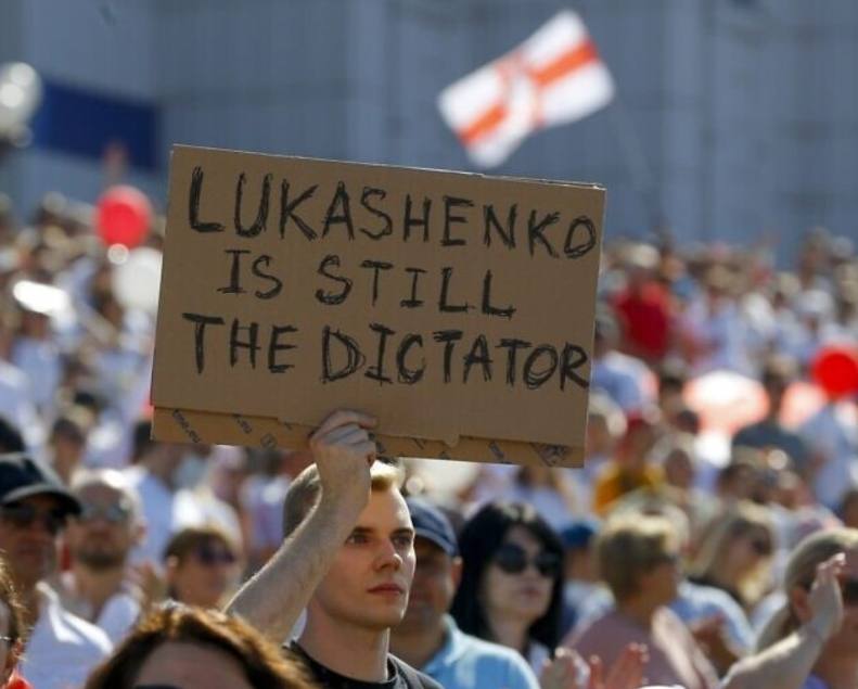 Na imagem há belarrussos em uma manifestação, aonde um jovem loiro segura um cartaz dizendo que Lukashenko continua sendo um ditador.