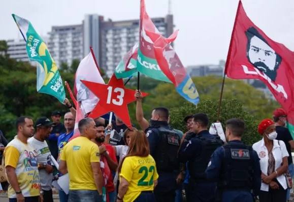 Petistas de vermelho, segurando bandeiras de apoio ao Lula e ao partido PT, e bolsonaristas segurando cartazes de apoio ao bolsonaro, usando camisas do Brasil, discutindo entre si.
