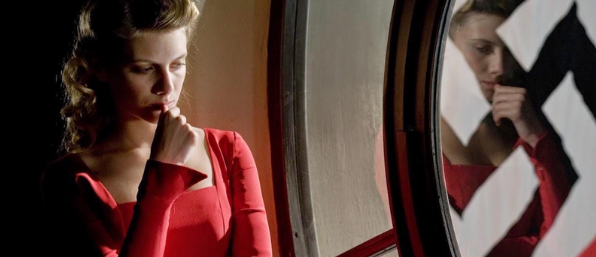 Na esquerda, mulher de vermelho olhando pensativa para um vidro a direita, o qual, pode-se ver seu reflexo e uma suástica.
